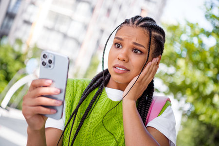 Young woman with braided hair takes a selfie in a sunny city day wearing a bright green knit sweater and pink backpack while listening to music on her phoneの写真素材