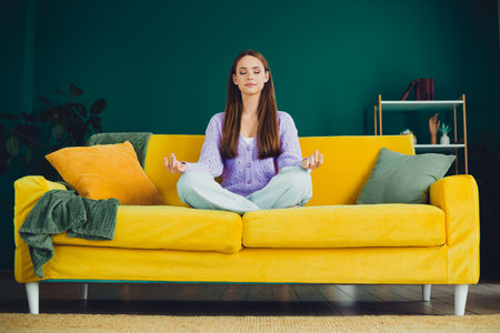 Meditative moment at home a young woman sits cross legged on a bright yellow sofa in a modern cozy living room sceneの写真素材