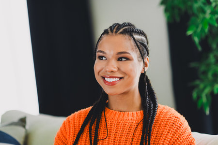 Smiling woman with braided hair wearing an orange knit sweater sits in a bright living room enjoying a relaxed day at homeの写真素材