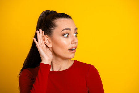 Young woman in red top listens with surprised expression against bright yellow background showcasing...