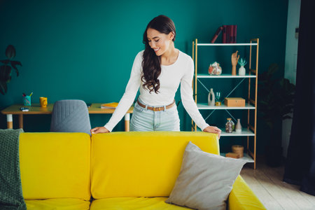Woman standing behind bright yellow sofa in a stylish living room with teal wall and decorative shelf creating a cozy home atmosphereの写真素材