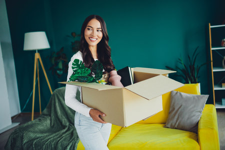 Moving day joy a young hispanic woman carries a cardboard box with plants into a bright modern living room at homeの写真素材