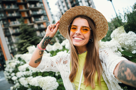 Cheerful young woman in sunhat poses for a selfie in a sunny city park with white flowers and stylish casual fashionの写真素材