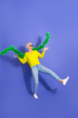 Joyful elderly woman leaps with green feather boa wearing yellow sweater and jeans against a purple backgroundの写真素材