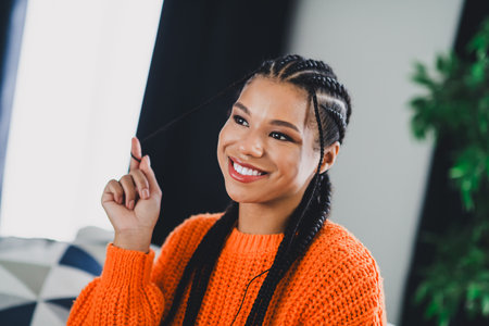 Bright living room scene with happy woman in orange sweater smiling and braided hairの写真素材