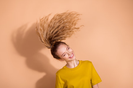 Vibrant young woman with flowing wavy hair against a beige background in casual yellow attire radiates happiness and styleの写真素材