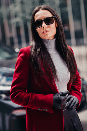 Stylish woman in a red coat and sunglass outdoors in a trendy urban setting on a sunny day.の写真素材