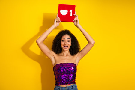 Young stylish woman with purple sequin top celebrates on yellow background holding a red sign with heartの写真素材