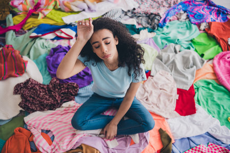High angle view full size photo of young girl sit pile stack clutter clothes hand touch head apartment indoorsの写真素材