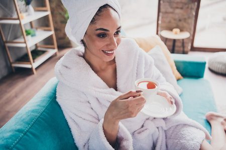 Photo portrait of pretty young girl sit sofa drink tea hold cup head wrapped towel dressed bath robe beauty routine home conceptの写真素材