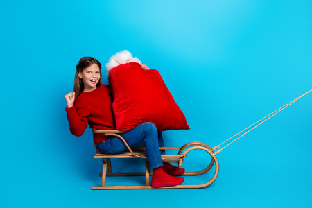 Joyful girl rides a wooden sleigh holding a bright red Santa sack against a vivid blue background during Christmas seasonの写真素材
