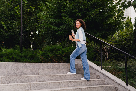 Casual young woman climbs stairs outdoors in an urban park holding coffee and phone smiling at the camera during a sunny dayの写真素材