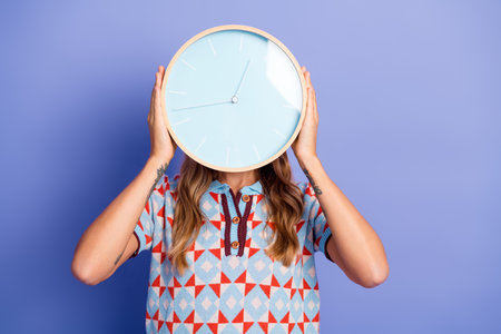 Young woman holds a large clock in front of a blue background wearing a geometric patterned shirt for a lifestyle fashion stock photoの写真素材