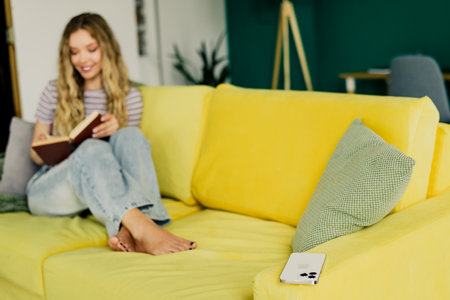 Bright day cozy reading moment at home as a woman sits on a yellow couch reading a book in a stylish living roomの写真素材