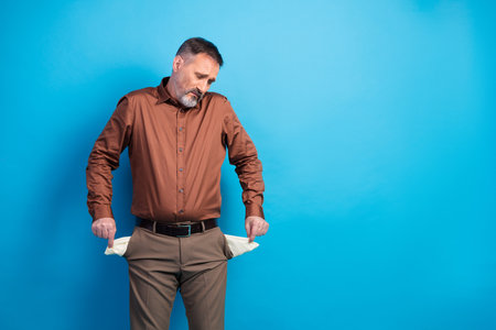 Businessman with empty pockets shows worry and honesty against blue background wearing brown shirt and beige pants during a professional studio shootの写真素材