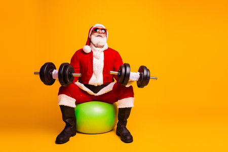 Santa lifts weights in a bright orange studio sitting on a green ball for holiday fitness and...
