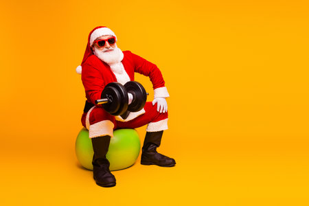 Santa lifts dumbbell in bright orange studio wearing red suit and hat smiling for festive fitness themeの写真素材