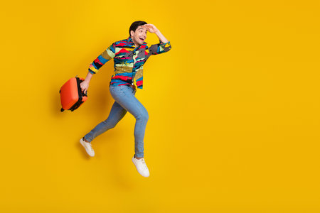 Lively young man in colorful shirt jumps with orange suitcase against bold yellow background for travel lifestyle and advertising useの写真素材