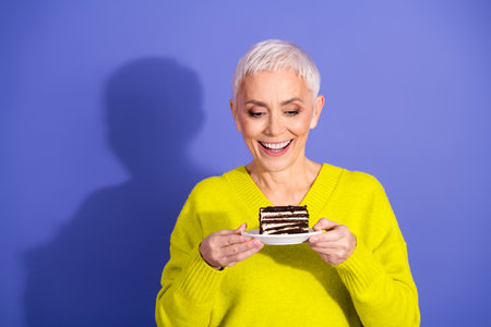 Happy elderly woman in bright yellow sweater smiles while holding a chocolate cake slice against a purple background for a warm lifestyle advertising imageの写真素材