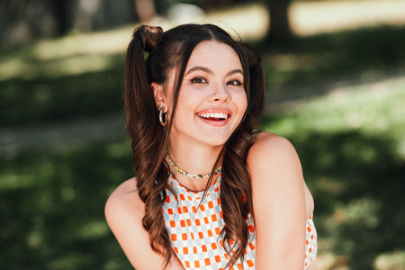 Young smiling brunette girl in park enjoying sunny day wearing checkered dress playful pigtails and carefree outdoor lifestyle vibeの写真素材