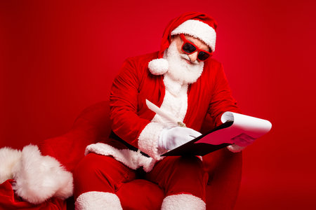 Santa reading script in red festive chair wearing costume with sunglass white beard as he acts for a humorous holiday sceneの写真素材
