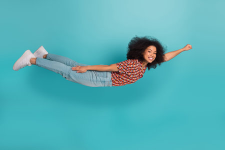 Young fashionable woman floating against blue background wearing printed shirt and jeans smiling with energy and joyの写真素材