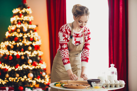 Fun holiday baking moment with a cheerful blonde woman in a cozy apron preparing dough cookies and treats by the lit Christmas treeの写真素材