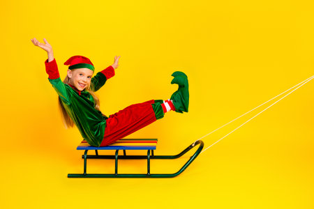 Cheerful child in an elf costume sitting on a sled with excitement on a vibrant yellow background, joyful holiday vibesの写真素材