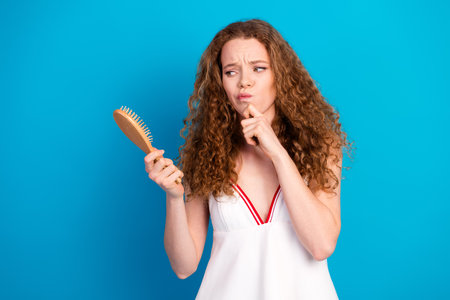 Young woman with curly brown hair holds a wooden brush and ponders beauty fashion and style against a blue backdropの写真素材