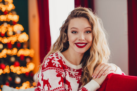 Playful woman in a red white Christmas sweater sits by a red sofa with a glowing Christmas tree in a cozy home setting for a festive holiday moodの写真素材