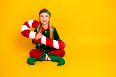 Adorable girl dressed as a festive elf holding a candy cane prop on bright yellow background, spreading holiday cheerの写真素材