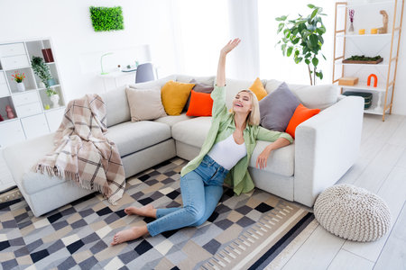 Full length photo of lovely young lady sit floor have fun enjoy weekend wear trendy green garment interior design spacious bright apartmentの写真素材