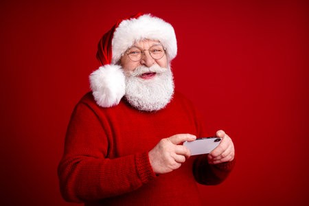 Santa plays on his phone in a red sweater and hat against a bold red backdrop for a joyful Christmas seasonの写真素材