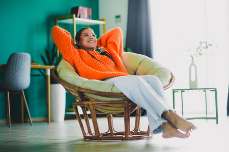 Relaxed woman lounging in a bright orange sweater in a stylish living room chair at home enjoying leisure time in a modern apartmentの写真素材