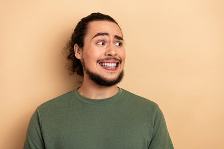 Young mixed race man with green shirt smiles awkwardly against a beige background looking to the...