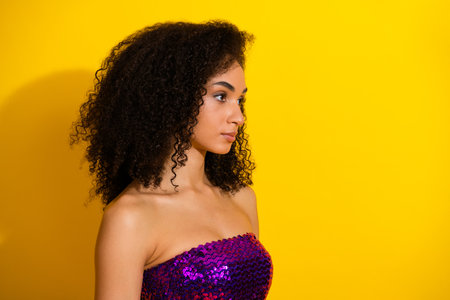 Young woman with curly hair wearing a purple sequin top stands against a vivid yellow background showcasing glamour and fashion confidenceの写真素材