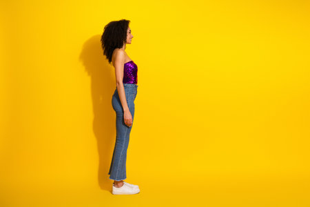Young stylish woman in a purple sequin top and jeans stands in profile against a bright yellow background for a fashionable casual lifestyle portraitの写真素材