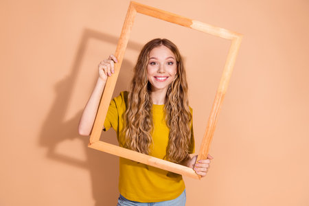Smiling young woman holding a wooden frame against a beige background with a casual and cheerful...