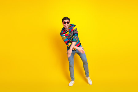 Young man in colorful shirt dances against a bright yellow background wearing sunglass and casual...