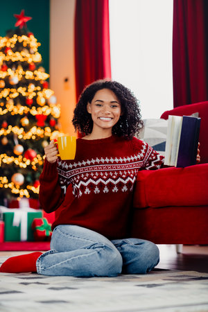 Cheerful woman in cozy sweater enjoys coffee and books beside a glowing Christmas tree at homeの写真素材
