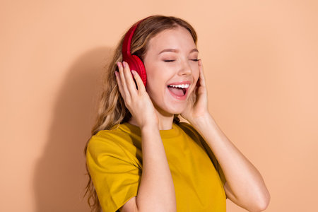 Young cheerful woman enjoying music with bright red headphones on pastel beige background, expressing happinessの写真素材