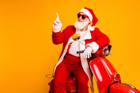 Santa on a bright yellow background points up while riding red scooter festive costume with hat beard for Christmas season holiday campaignsの写真素材