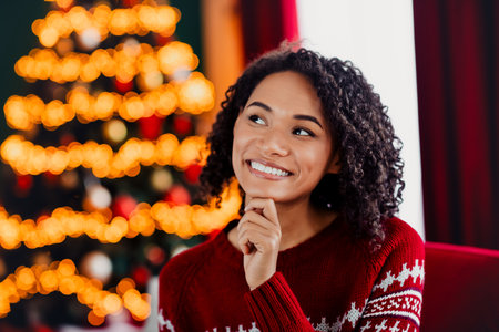 Cheerful woman in Christmas sweater thinking by warm lit tree dreaming of merry moments at home this holiday seasonの写真素材