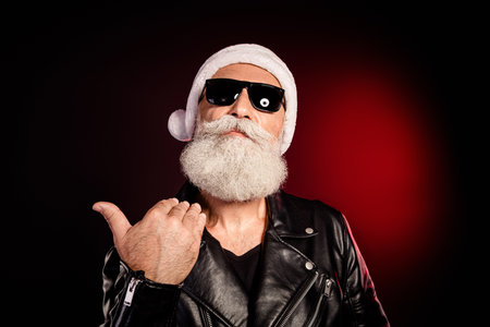 Santa rocker in leather jacket with white beard and hat gives thumbs up against dark red background...