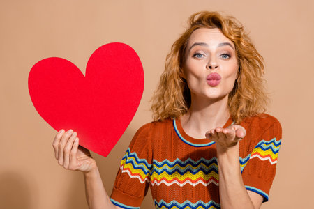 Photo portrait of lovely young lady hold big red heart postcard kiss dressed stylish striped garment isolated on beige color backgroundの写真素材