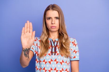 Young woman with blonde hair stands against a blue background raising her hand in a stop gesture and looking serious and confidentの写真素材
