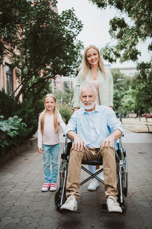 Cheerful family day outdoors with grandpa in wheelchair being cared for by daughter and wife in a sunny parkの写真素材