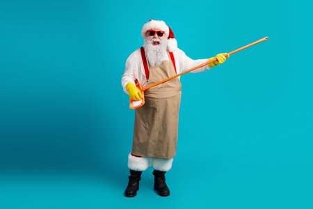Santa in funny cleaning outfit with orange mop on bright blue studio backdrop celebrating Christmas season and holiday cheer festive spiritの写真素材