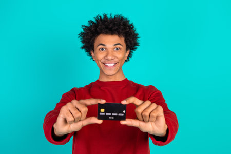 Young man holding a credit card toward the camera smiling in a red shirt against a teal blue background perfect for shopping fashion lifestyleの写真素材