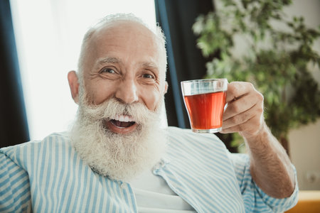 Candid senior man with glass of juice in bright home interior enjoying a cheerful relaxed momentの写真素材